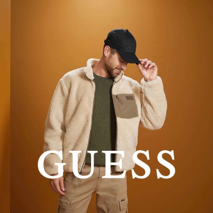 Mężczyzna w beżowej polarowej kurtce GUESS i czapce z daszkiem, na ciepłym brązowym tle.