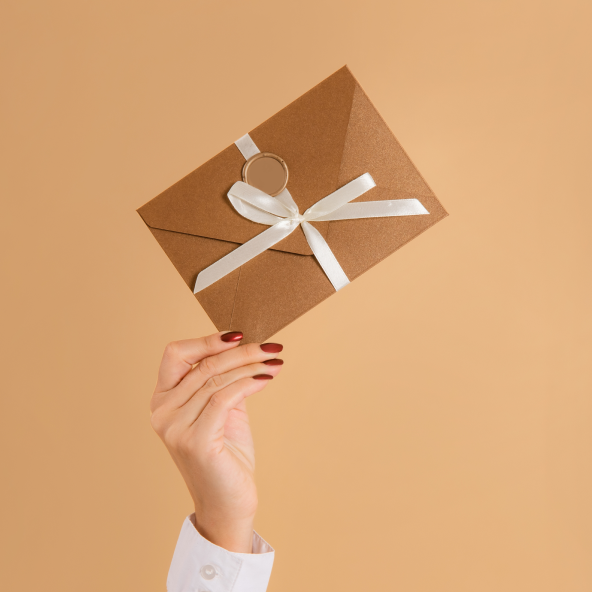 ręka trzymająca beżową kopertę przewiązaną wstążką, na beżowym tle