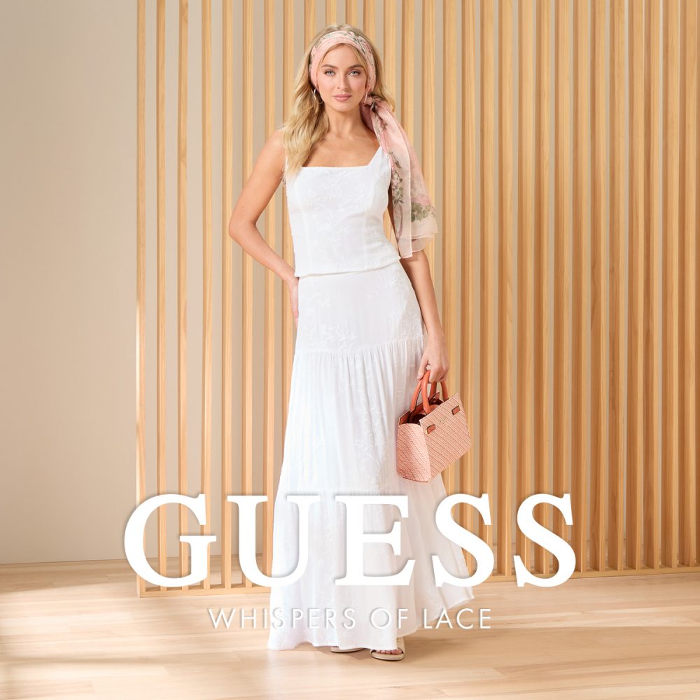 Modelka w białym komplecie Guess Whispers of Lace, składającym się z topu i długiej spódnicy, z torebką i apaszką na głowie, na tle drewnianych paneli.
