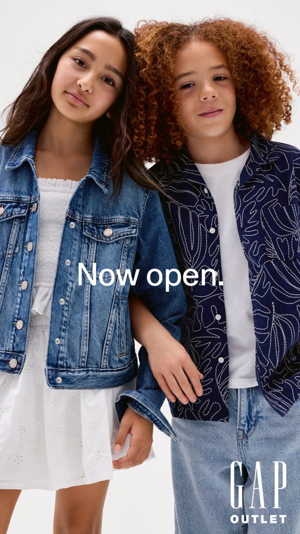 Dwoje dzieci w ubraniach GAP, dziewczynka w jeansowej kurtce i białej sukience, chłopiec w jeansach i koszuli we wzory, z napisem 'Now open.' i logo GAP Outlet.