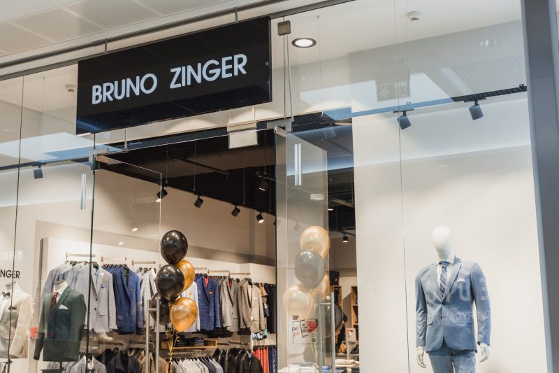 Wejście do sklepu Bruno Zinger z manekinem w garniturze i balonami, widoczne ubrania na wieszakach.