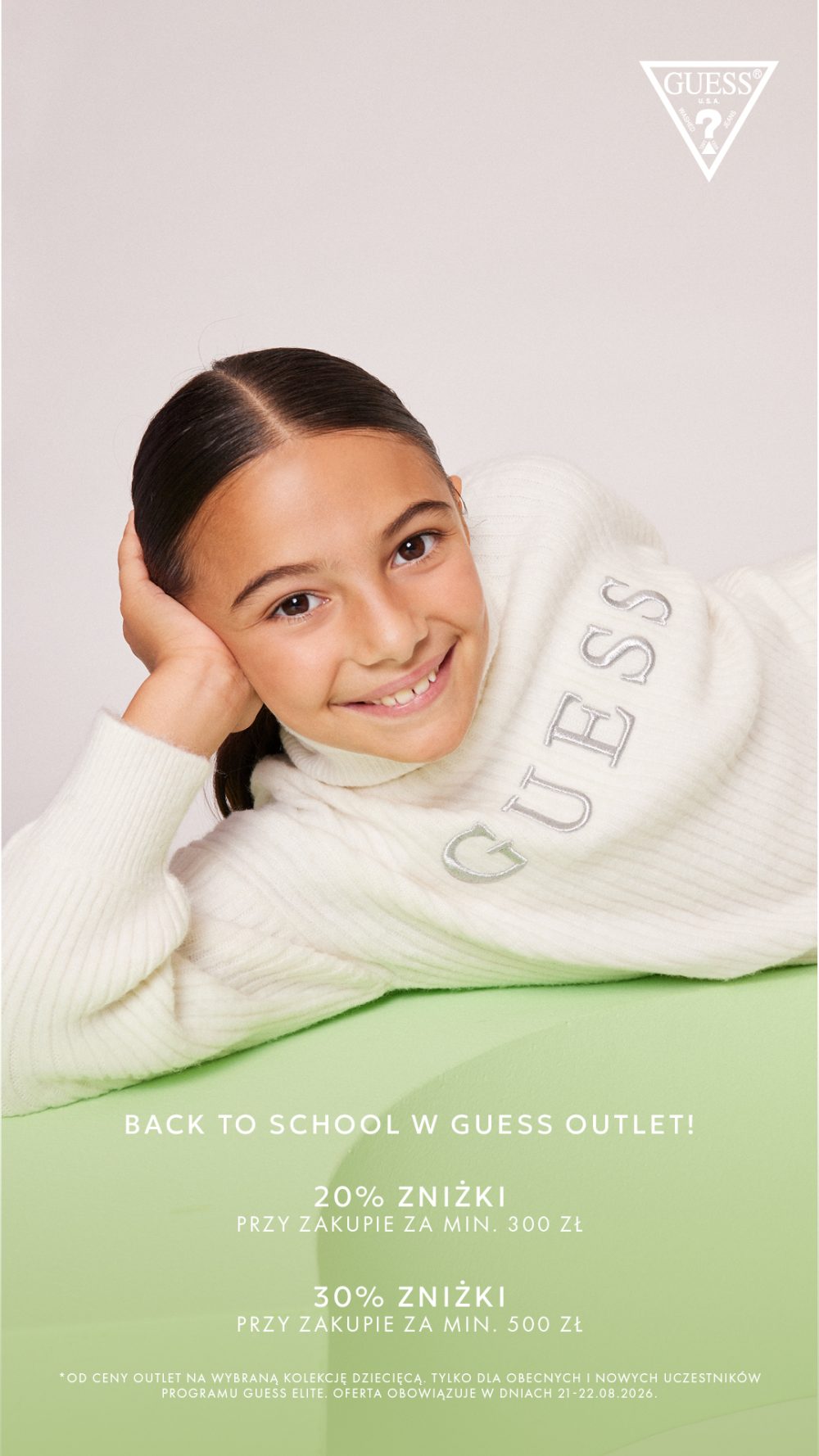 Uśmiechnięta dziewczynka w białym swetrze Guess, leżąca na zielonym tle, promująca zniżki Back to School.