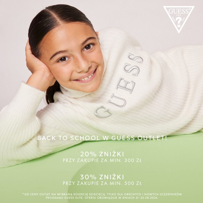 Uśmiechnięta dziewczynka w białym swetrze Guess, leżąca na zielonym tle, promująca zniżki Back to School.