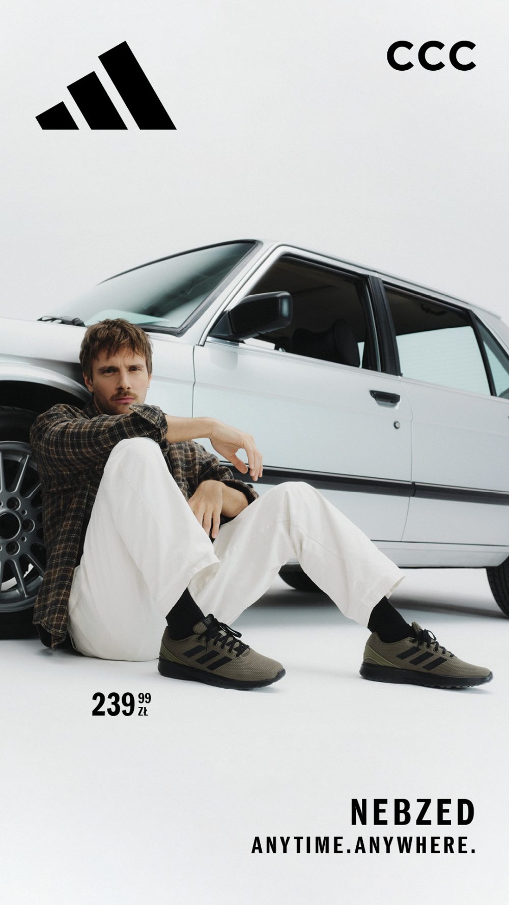 Maciej Musiał siedzi obok srebrnego samochodu, ubrany w białe spodnie, koszulę w kratę i zielone buty adidas Nebzed. W tle logo adidas i CCC.
